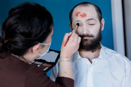 Girlfriend applying professional eye makeup on man to make him look dead and diseased for halloween, dressing him as zombie. Woman applying foundation for pale face and scars cosmetics on boyfriendの写真素材