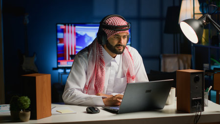 Arab employee with wireless headphones working from living room office, sending emails. Muslim worker doing internet research using laptop while listening to music to make time pass fasterの写真素材