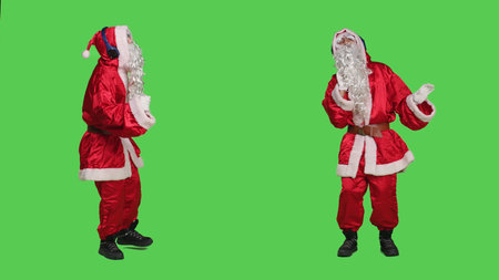 Man in red suit having fun with audio, listening to music on headphones to celebrate winter seasonal holiday. Santa with white beard dancing and enjoying songs over greenscreen backdrop.の写真素材