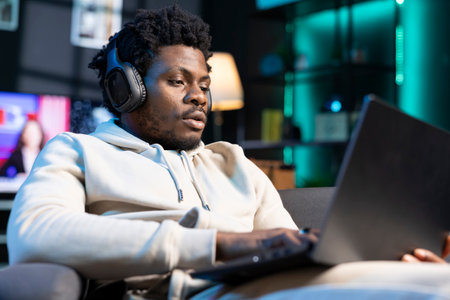African american man relaxing at home by watching TV series on laptop using headphones. Carefree person enjoying leisure time in RGB lit living room at night, consuming entertainment media contentの写真素材