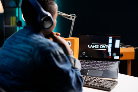 Black woman sitting and looking at her laptop screen displaying game over. Over the shoulder shot of african american female gamer gazing at personal computer after losing video game.の写真素材