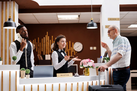 Friendly concierge checking senior man in at the main desk of a luxury hotel. While luggage is close in the lounge area, a pleasant grin from a female employee helps an elderly male tour guide.の写真素材