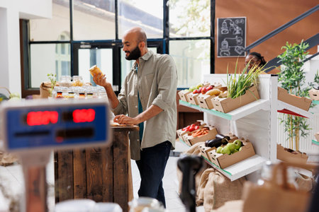 Male customer holds and admires a glass container filled with an organic food product in eco friendly grocery store. Middle eastern man grasping and looking at a jar of natural honey.の写真素材