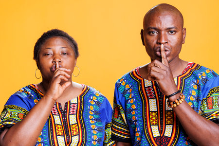 African american man and woman holding forefinger on lips and asking to keep secret portrait. Couple making shh gesture, showing silence signal and looking at camera with serious expressionの写真素材
