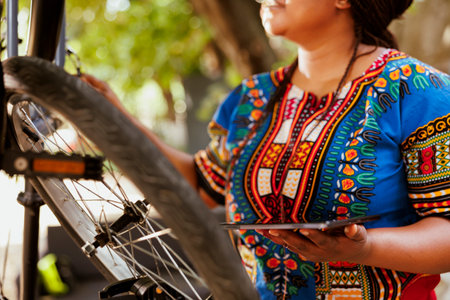 Detailed shot of healthy sports-loving black woman searching on phone tablet for bike summer maintenance. Image showing close-up view of african american female holding smart device while maintaining bicycle.の写真素材