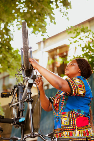 Young african american woman engrossed in repairing modern bicycle with professional work tool. Active determined female cyclist confidently repairing bike chain and inspecting tire for outdoor leisure cycling.の写真素材