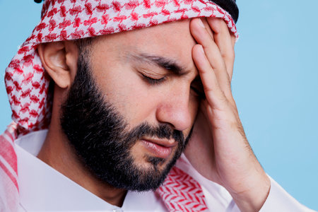 Arab man with exhausted expression holding forehead while suffering from migraine symptom. Muslim person dressed in traditional islamic clothes and headdress having headacheの写真素材