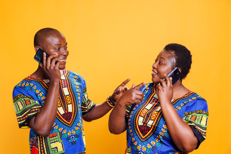 Smiling african american man and woman looking at each other and gesticulating while chatting on smartphone. Cheerful couple speaking on mobile phone, having fun discussionの写真素材