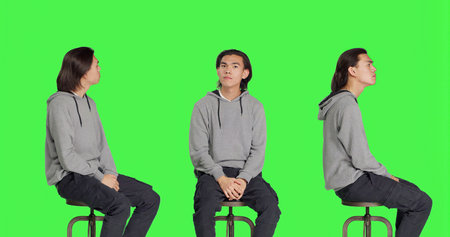 Young man sitting on chair and waiting for something in studio, looking at watch and searching for a thing against greenscreen backdrop. Male model with natural impatient expressions.の写真素材
