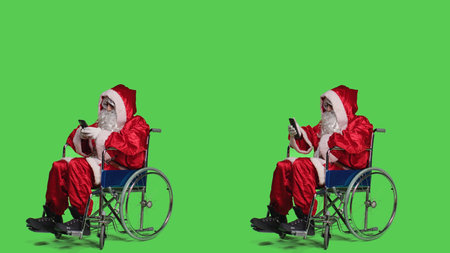 Santa texting and being on videocall dealing with chronic impairment, using smartphone against full body greenscreen. Young man saint nick talks on online teleconference meeting.の写真素材