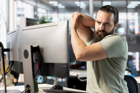 Startup employee suffering arm pain from bad posture on office chair while doing research for project. Man feeling muscle strain, stretching while looking at computer in workspace, solving tasksの写真素材