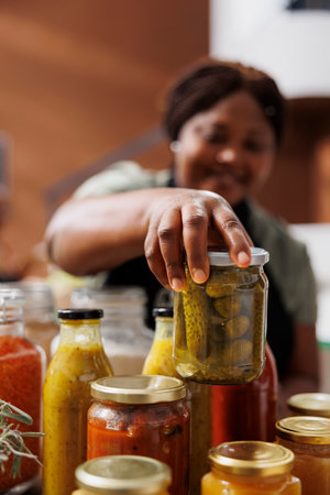 African American woman, dressed as a shopkeeper, sells preserved food in glass jars. Close up of black female vendor holding and arranging container of pickles in eco friendly grocery store.の写真素材