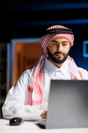Image shows an Arabian man working at a desk, using a laptop and wireless technology. Muslim male embraces modern tech for his entrepreneurial endeavors by utilizing his personal computer.の写真素材