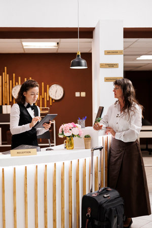 Elderly lady stands at hotel reception, where professional concierge assists with reservation. Senior woman completes check-in process with digital document on tablet. Suitcase is placed near counter.の写真素材