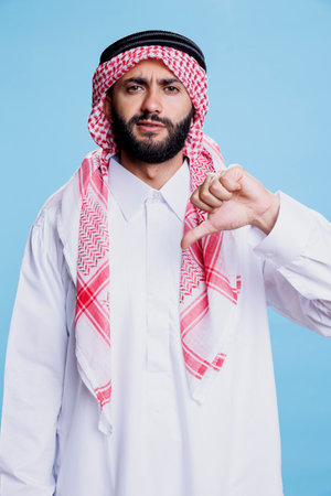 Arab man in traditional robe and headdress showing thumb down studio portrait. Confident muslim person wearing white thobe showcasing disapproval gesture and looking at cameraの写真素材