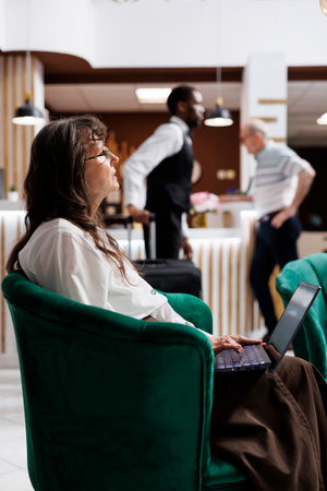 Side-view shot of elderly female customer sitting in hotel lounge area, browsing the internet on her laptop. Close-up of retired senior woman using her personal computer for online check-in.の写真素材