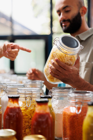 Male customer holds a glass jar filled with an organic food product in an eco friendly store. Middle eastern man is seen listening to advice of the vendor while grasping a plastic free container.の写真素材