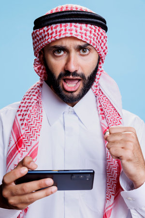 Excited muslim man wearing traditional clothes playing mobile game and celebrating success studio portrait. Happy arab internet gamer holding smartphone and looking at camera with clenched fistの写真素材