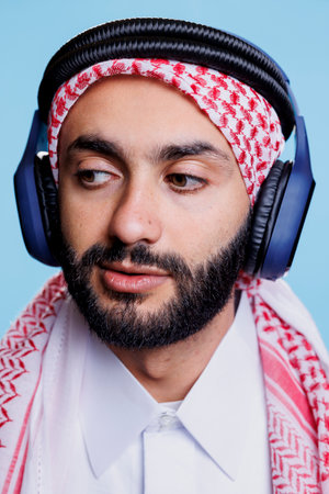 Man wearing checkered traditional muslim headscarf listening to music in headphones and looking away closeup. Young arab in traditional clothes enjoying song in wireless earphonesの写真素材