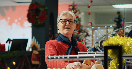 Portrait of elderly woman browsing through clothes in Christmas decorated clothing store during...