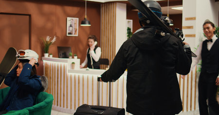 Man asking staff about extreme sports while he holds skis gear in alpine resort entrance, tourism industry during winter. Hotel guest ready to have fun on snowy slopes for leisure. Handheld shot.の写真素材