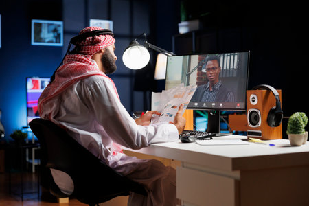 Image showcasing a Muslim guy grasping paperwork while having a conversation with his African-American coworker. Having a video call on desktop computer are two young businessmen.の写真素材