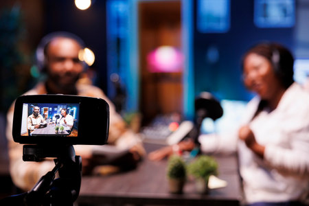 During live talk show broadcast on streaming platforms, cohosts use a professional camera for recording. African vloggers seen in blurry background using modern recording equipment to make a vlog.の写真素材