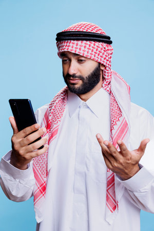 Arab man in traditional clothes engaging in virtual conversation on mobile phone. Person wearing muslim headscarf and thobe speaking in videocall and making gesture with handの写真素材