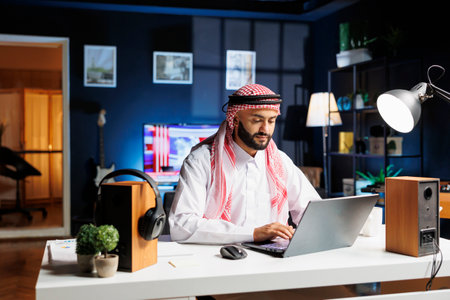 Focused Arab entrepreneur works efficiently typing on a laptop and browsing the internet. Dressed in traditional clothing, a male Muslim person engrossed in his work, embracing technology.の写真素材