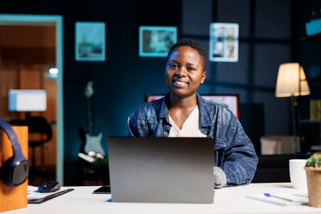 Portrait shot of smiling black woman working from home by using her personal computer. African american entrepreneur looking at camera and utilizing a digital laptop for research.の写真素材