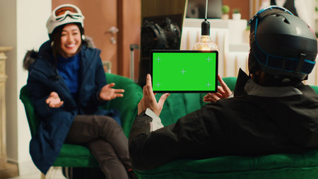 Young man uses tablet with greenscreen display in ski resort lounge area, holding digital gadget displaying blank chroma key mockup template. Couple talking about extreme winter sports.の写真素材