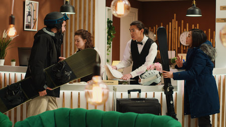 Diverse people ask staff about skiing, travelling at mountain winter resort for winter sports on snowy slopes. Man and woman arriving at hotel front desk to discuss about room services, carry gear.の写真素材