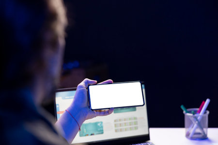 Male individual holding a smartphone horizontally with an isolated white screen. A person is using a laptop and grasping a cell phone that has a blank copyspace mockup template.の写真素材