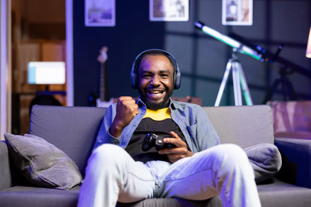 Cheerful man wearing headphones, celebrating after victory in videogame, neon lit apartment. Person...