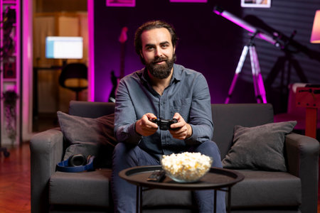 Portrait of smiling man playing shooter videogame in neon lit room, holding controller. Gamer participating in internet esports competition using professional gamepad and eating popcornの写真素材