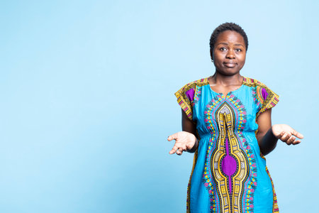 African american woman being clueless and unsure over blue background, dressed in traditional ethnic attire in studio. Young doubtful person acting uncertain and confused.の写真素材