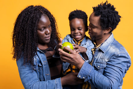 Adorable small child looking at an apple against yellow background, posing with his mother and...