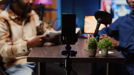 Focus on smartphone on tripod used by podcast host in blurry background to record guest telling...