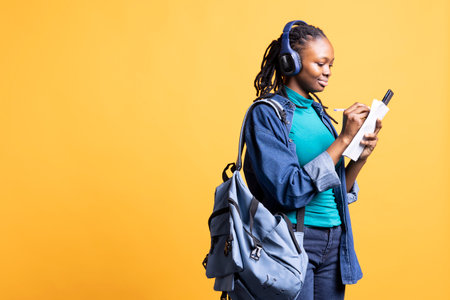 Student writing homework, doing research online on smartphone and listening music, studio background. Girl preparing for school exam, taking notes and enjoying songs on headphonesの写真素材