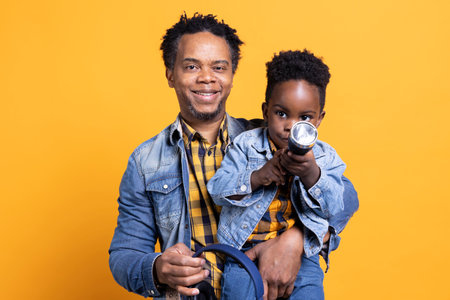 Smiling african american father is carrying his son against yellow background, curious little boy playing with a black flashlight in front of the camera. Cheerful dad bonding with his kid.の写真素材