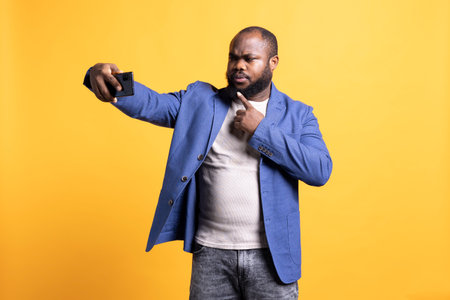 Confident african american man using smartphone to take selfies, pointing finger to chin. Person taking photos using mobile phone selfie camera, studio backgroundの写真素材