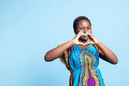 Young person showing romantic gesture with heart shape sign, wearing traditional attire over blue background. African american girl expressing lovely emotions and her true feelings on camera.の写真素材
