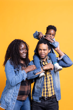 African american dad carrying his boy on his shoulders in studio, cute small kid posing on camera with a hand. Cheerful loving mother and father bonding with their son.の写真素材