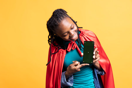 Joyous girl portraying superhero with cape showcasing green screen phone, isolated over studio...