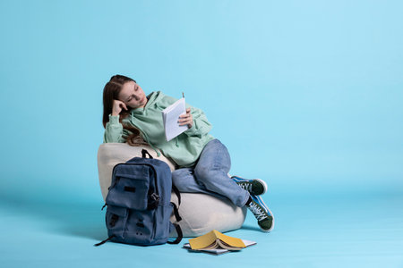 Portrait of pensive girl sitting on beanbag, doing head propped by arm thinking gesture, studio background. Contemplative schoolgirl with notepad in arms reading what she wrote on homework so farの写真素材