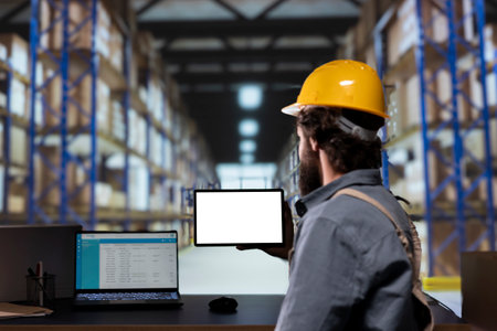 Warehouse manager looking at display with white layout on tablet, working in storage room depot with...