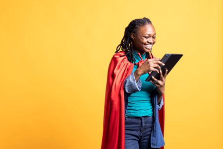 Joyous girl portraying superhero with red cape using tablet, isolated over studio background. Joyful teenager posing as hero in costume typing on digital device touchscreenの写真素材