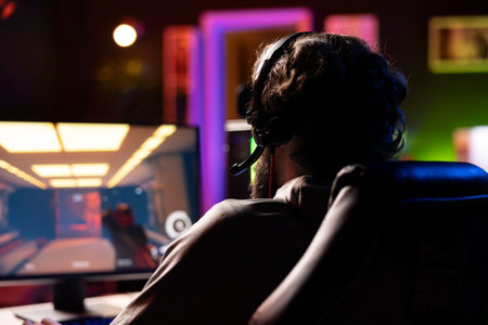 Man in dark living room playing engaging video games on gaming PC at computer desk, chilling after work. Close up of gamer wearing headphones, talking with friends in online multiplayer shooterの写真素材