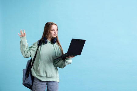 Happy woman saluting friends during videoconference meeting using laptop, studio backdrop. Girl waving hand, greeting mates during internet videocall on notebook, camera Aの写真素材