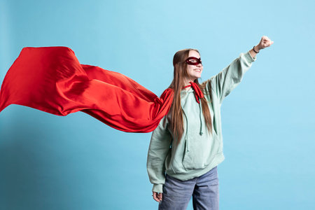 Cheerful girl wearing superhero costume and mask for Halloween with cape fluttering, studio background. Portrait of jolly teenager smiling while dressed as comic book hero, pretending to flyの写真素材